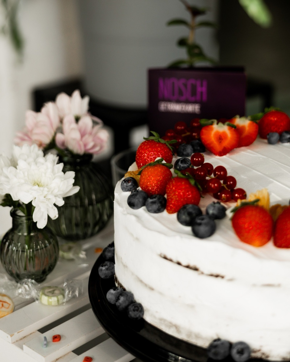 Vegane Erdbeertorte mit Erdbeeren, Blaubeeren und Johannisbeeren, daneben Blumen in Glasvasen