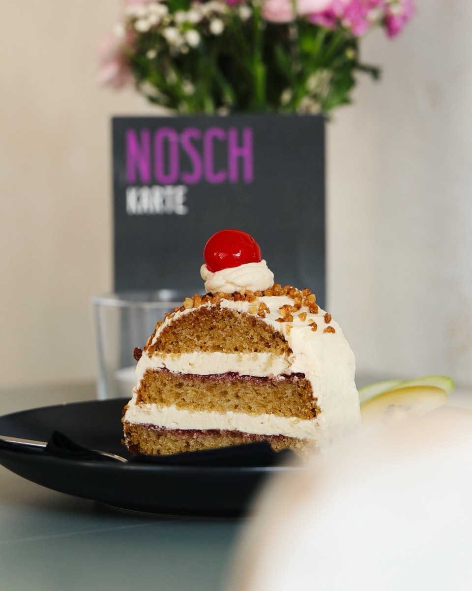Ein Stück veganer Frankfurter Kranz mit Krokant und Kirsche, dahinter die NOSCH-Karte und Blumen