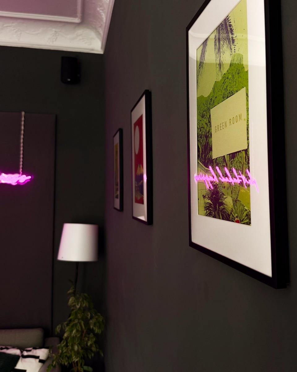 Gerahmte Poster aus der Reihe „Leipziger Collagen“ von Atelier Zündwerk, darunter „Green Room“, an einer dunklen Wand im NOSCH, in der Scheibe spiegelt sich der pinke Neon-Schriftzug „plant-baked happiness“
