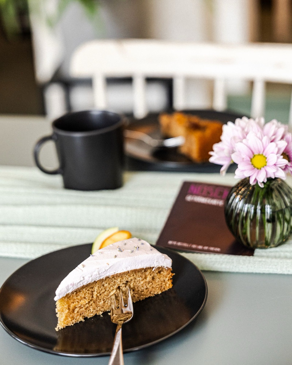 Ein Stück veganer London Fog Cake mit zartrosa Creme und Lavendelblüten, dahinter eine Tasse und Blumen im NOSCH