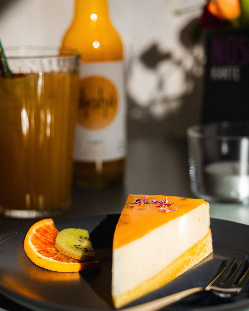 Ein Stück veganer Maracuja-Cheesecake mit Orange und Kiwi, dahinter ein Glas Saft in der Sonne